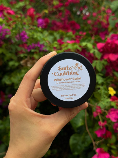 Hand holding a container of Sudz Cauldron Wildflower Flores de Paz Balm against a floral background