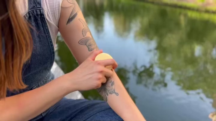 Person gliding a solid lotion bar over their arm beside a calm lake, sunlight reflecting on the water.