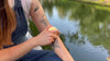 Person gliding a solid lotion bar over their arm beside a calm lake, sunlight reflecting on the water.